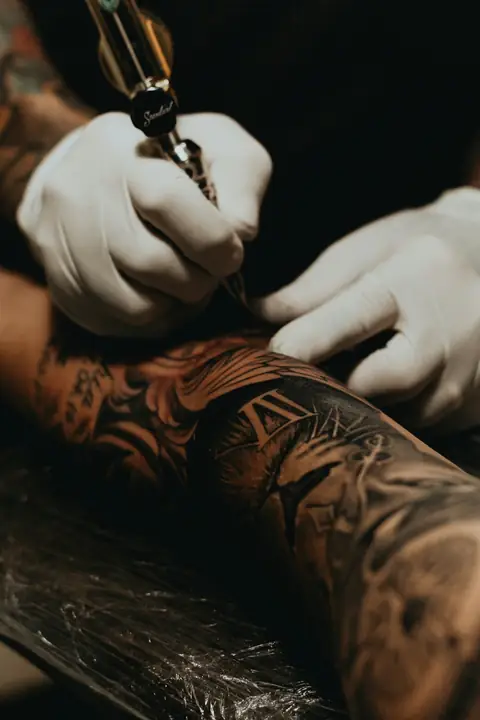 Gloved hands tattooing a heavily tattooed forearm with a rotary pen