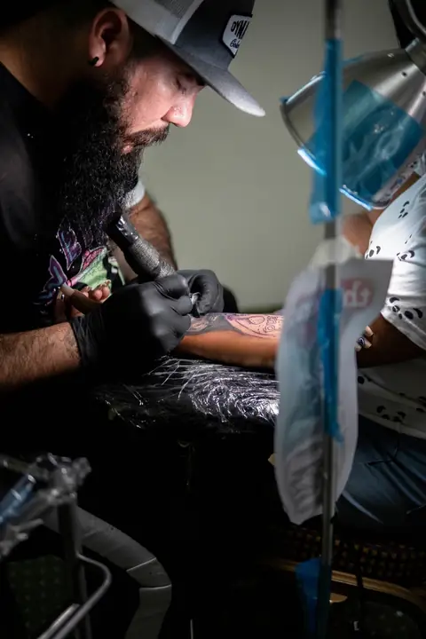 Tattoo artist in a cap working on a client’s forearm